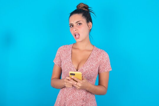 Photo of astonished crazy beautiful brunette woman wearing floral dress over blue background hold smartphone dislike feedback concept