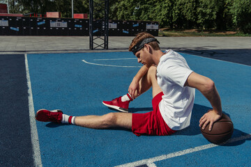 Fototapeta na wymiar basketball player in red shorts and sneakers sitting on court near ball