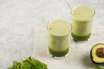 A glass of green healthy detox smoothie with spinach, matcha powder and a glass straw, fresh spinach and avocado on marble board on grey background