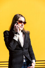 Executive entrepreneur woman, wearing black jacket and wool sweater and sunglasses, on yellow background in the street, with copy space, talking on the mobile. Entrepreneur woman concept.