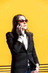 Executive entrepreneur woman, wearing black jacket and wool sweater and sunglasses, on yellow background in the street, with copy space, talking on the mobile. Entrepreneur woman concept.