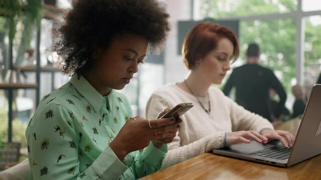 People using cellphone and laptop at coffee shop. Two young women staring at screens. Girl looking at computer browsing internet inside cafe restaurant