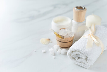 Obraz na płótnie Canvas Spa composition with towel, small wooden bowl with coarse sea salt, vial of oil, candle, skin cream, sea shell on the light marble background. Copy space. Top view