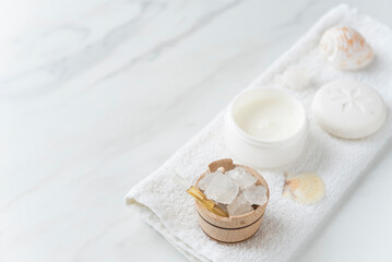 Fototapeta na wymiar Spa composition with small wooden bowl full of coarse sea salt, skin cream, soap, sea shells on the white towel on the light marble background. Copy space. Flat lay