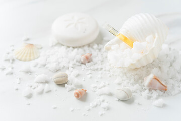Obraz na płótnie Canvas Beautiful white sea shell full of coarse salt and vial of oil, soap and scattered salt, small sea shells and stones on the light marble background. Spa. Flat lay