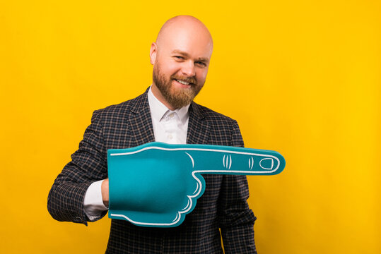 Bearded man is holding a fan blue glove while he is pointing away at a free space over yellow wall - Powered by Adobe