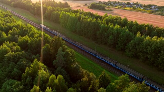 Passenger train on railway from forest, aerial view. Train with passenger cars rides along forests on sunset. Drone view of an electric passenger locomotive with wagons in moving on railroad.