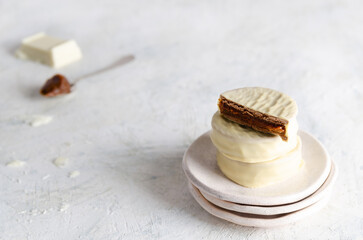 White chocolate alfajores on white plates, a piece of white chocolate and a spoonful of dulce de leche, on light background. 