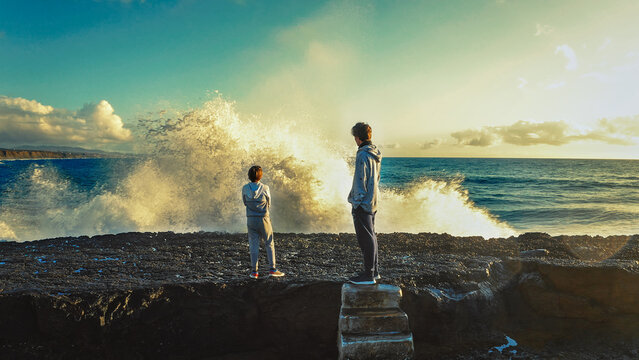 Boys look at the big waves crashing on the pier. The brothers contemplate the splashes of waves crashing on the rocky shore during sunset. Children stand and looking on the ocean. Freedom and travel