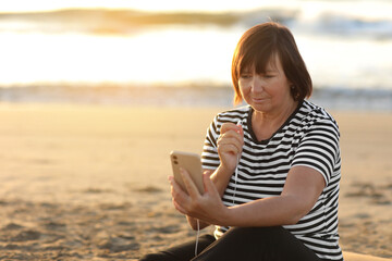 Smiling mature caucasian woman listening music in headphones looking at camera relaxing and resting on sandy beach by the sea at sunrise. Relaxation concept