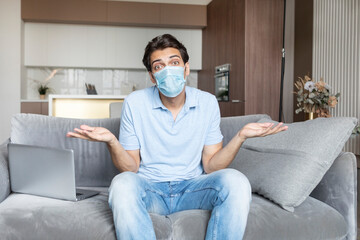 Unhappy man wearing medical mask and sitting on the couch and depressing about new covid-19 restrictions. He doesn't know what to do