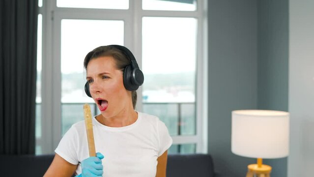 Woman in headphones cleans the house and have fun singing with a broom like a star. Slow motion