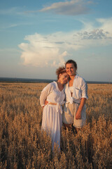 a mother and an adult daughter in white clothes walk through an oat field at sunset