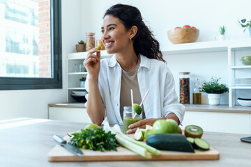 Pretty woman eating a piece of cucumber while drinking fruit detox juice in the kitchen at home.