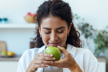 Beautiful young woman eating a green apple in the kitchen at home.