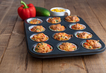 Twelve vegetable muffins in a baking form with pepper, corn and zucchini in the background, focus on foreground
