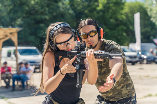 Shooting range concept. Private shooting lessons with an instructor. Medium outdoor shot of a powerful long-haired woman wearing protective equipment at aiming at the target with black riffle with