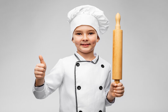 cooking, culinary and profession concept - happy smiling little boy in chef's toque and jacket with rolling pin showing thumbs up over grey background