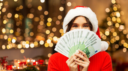 christmas, holidays and finance concept - happy woman in santa helper hat hiding behind euro money...