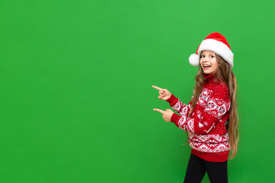 A little girl in a Santa Claus hat and a festive sweater points to your advertisement and smiles broadly on a green isolated background. Copy space.