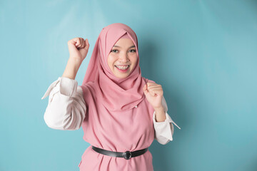 A young Asian Muslim woman with a happy successful expression wearing pink hijab isolated by blue background