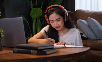 Pretty young woman in headphones writing important information on notebook during online lecture via laptop.