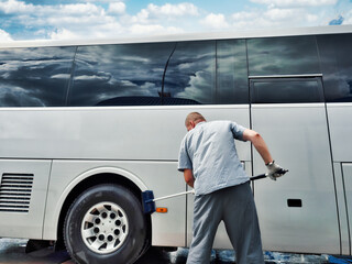 driver washes large bus with brush in open parking lot on summer day. Caucasian man prepares bus to transport passengers. Authentic scene workflow.