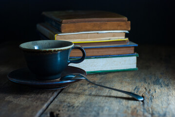 Coffee cup with saucer and coffee spoon with a pile of old books on a rustic wooden floor.