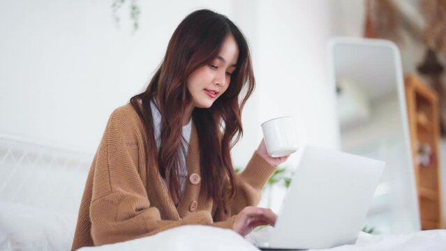 Happy beautiful young asian woman working on computer laptop and sitting on bed in house