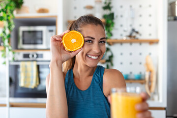 Beautiful young woman drinking fresh orange juice in kitchen. Healthy diet. Happy young woman with glass of juice and orange at table in kitchen.