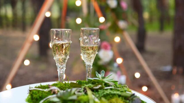 Wine glasses, wedding rings bedding of grass and moss on white table next to bohemian tipi arch decorated in boho style with flowers and candles and white chairs for guests.