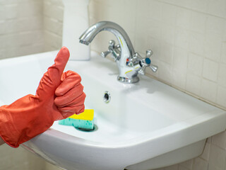 Cleaning bathroom sink and faucet with detergent in orange rubber gloves with green sponge. Housework, cleaning, hygiene home concept. Hand in protective glove showing like in front of bathroom sink.