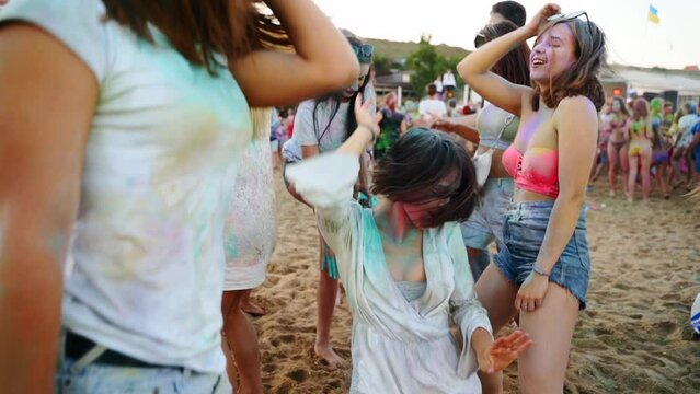 Young women smeared in colorful powder dance on sand at Holi festival on beach in slow motion. Outdoor party. Hindu holiday of colors, unity, love. End of lockdown, covid pandemic, restrictions.