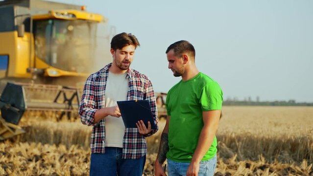 Farmers stand in wheat field, discuss harvesting plan and yield. Agronomist, logistics agent with touch tablet pc negotiate while harvester passing by. Rancher, landlord sign agreement on grain supply