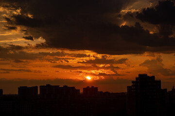 Sunset with gold, red, yellow and orange sunlight coming through the clouds, in a city among tall black houses. Horizontal photograph. Nature. Architecture.