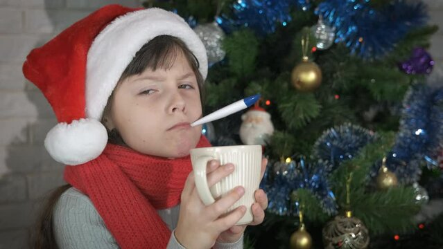 Sick Christmas with child with temperature. A sick younger with temperature stay in her Santa hat with a cup of tea by the Christmas tree.