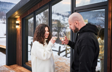 Smiling woman and man clinking glasses of wine and celebrating event together on winter day. Couple in love toasting enjoying alcoholic drink while standing outside scandinavian house barnhouse.