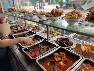 Padang food stalls share a variety of buffet menus with vegetables, chicken curry sauce, beef, eggs, tofu, tempeh. the waiter prepares the food. Menu in glass display case. Asia and asian food. Pile.