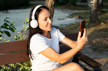 35 yeaers old woman during online video call. She is sittin on the bench in headptones with her smartphone.