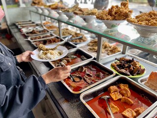Padang food stalls share a variety of buffet menus with vegetables, chicken curry sauce, beef, eggs, tofu, tempeh. the waiter prepares the arrangement of the food. Menu in glass display case.