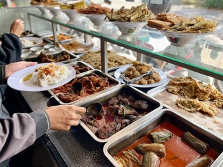 Padang food stalls share a variety of buffet menus with vegetables, chicken curry sauce, beef, eggs, tofu, tempeh. the waiter prepares the arrangement of the food. Menu in glass display case.