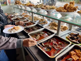 Padang food stalls share a variety of buffet menus with vegetables, chicken curry sauce, beef, eggs, tofu, tempeh. the waiter prepares the food. Menu in glass display case. Asia and asian food. Pile.