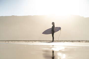 Beautiful surfer girl at the beach standing with her surfboard at sunset time. Female surfer