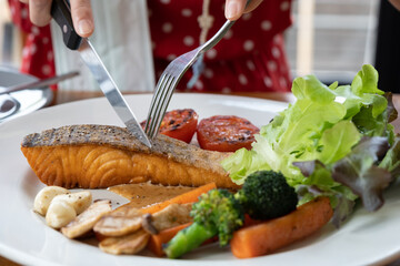 Woman's hand is using a knife to cut Salmon steak in a white plate on the dining table in the restaurant