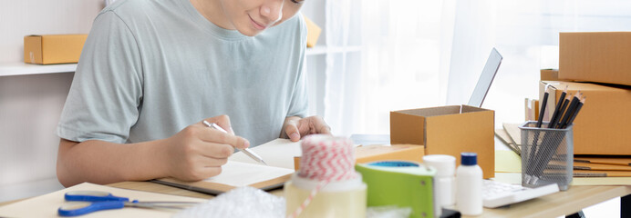 Young Asian man is writing down the customer's details and addresses on the notebook or box in order to prepare for shipping according to the information, Packing box, Sell online, freelance working.