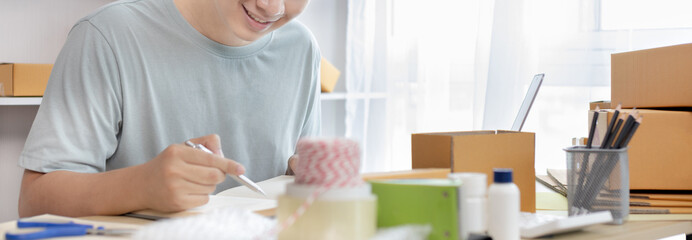 Young Asian man is writing down the customer's details and addresses on the notebook or box in order to prepare for shipping according to the information, Packing box, Sell online, freelance working.