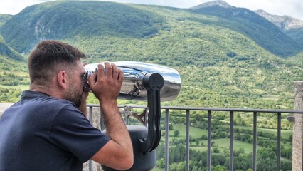 A young man looks through large street binoculars at beautiful landscapes. male tourist using a viewing machine to observe the panorama of nature with sea, forest and mountains
