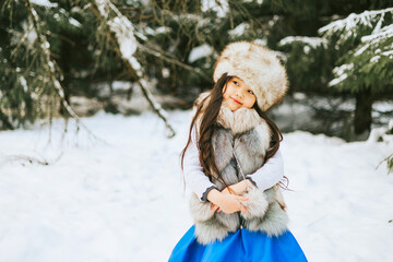 portrait of beautiful cute little Asian girl in fur coat and fur hat standing in snowy forest in winter, winter holidays and having fun, Christmas vacation