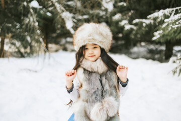 portrait of beautiful cute little Asian girl in fur coat and fur hat standing in snowy forest in winter, winter holidays and having fun, Christmas vacation