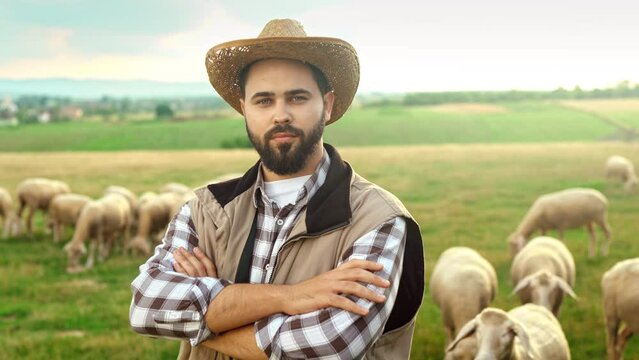 Portrait shot of handsome Caucasian young man in motley shirt and hat standing in field on summer with crossed hands and smiling to camera. Happy male shepherd posing at grassland with sheep flock.